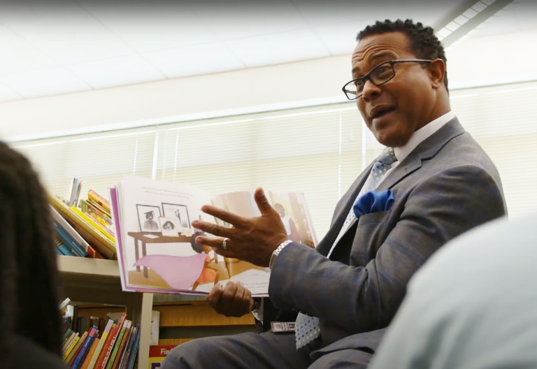African American man in a suit reading a children's book to a group of kids.