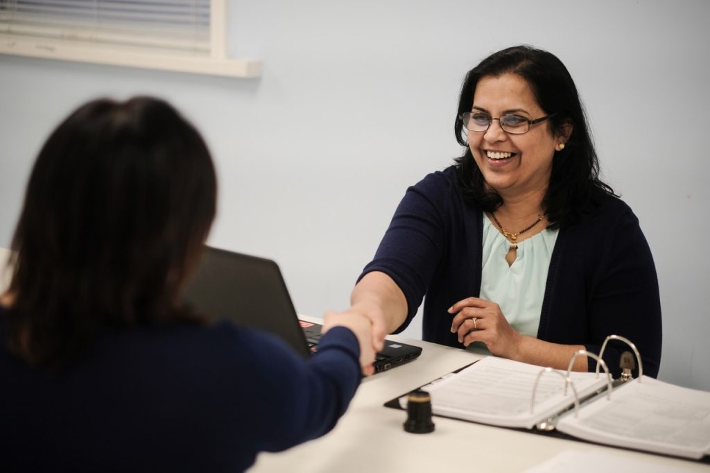 Smiling hispanic woman shaking hands with another woman across a desk.