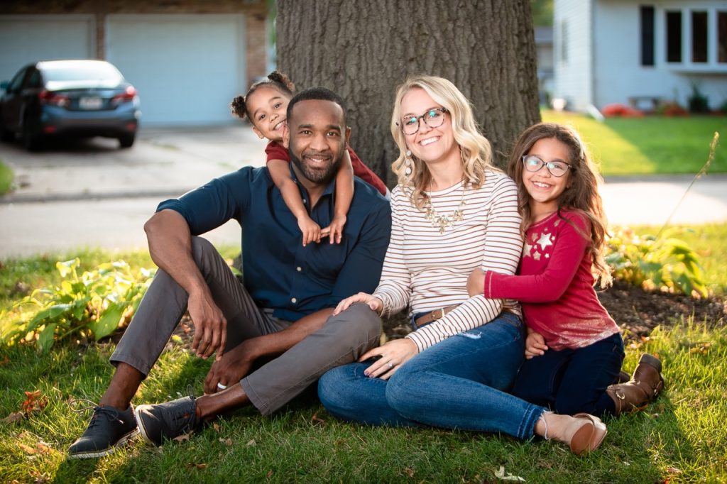 Family of husband, wife, and two kids sitting in the grass next to a tree.