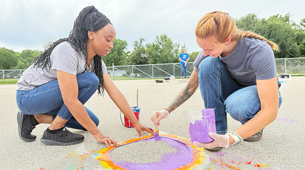 two women kneeling down outside painting a blacktop with bright colors